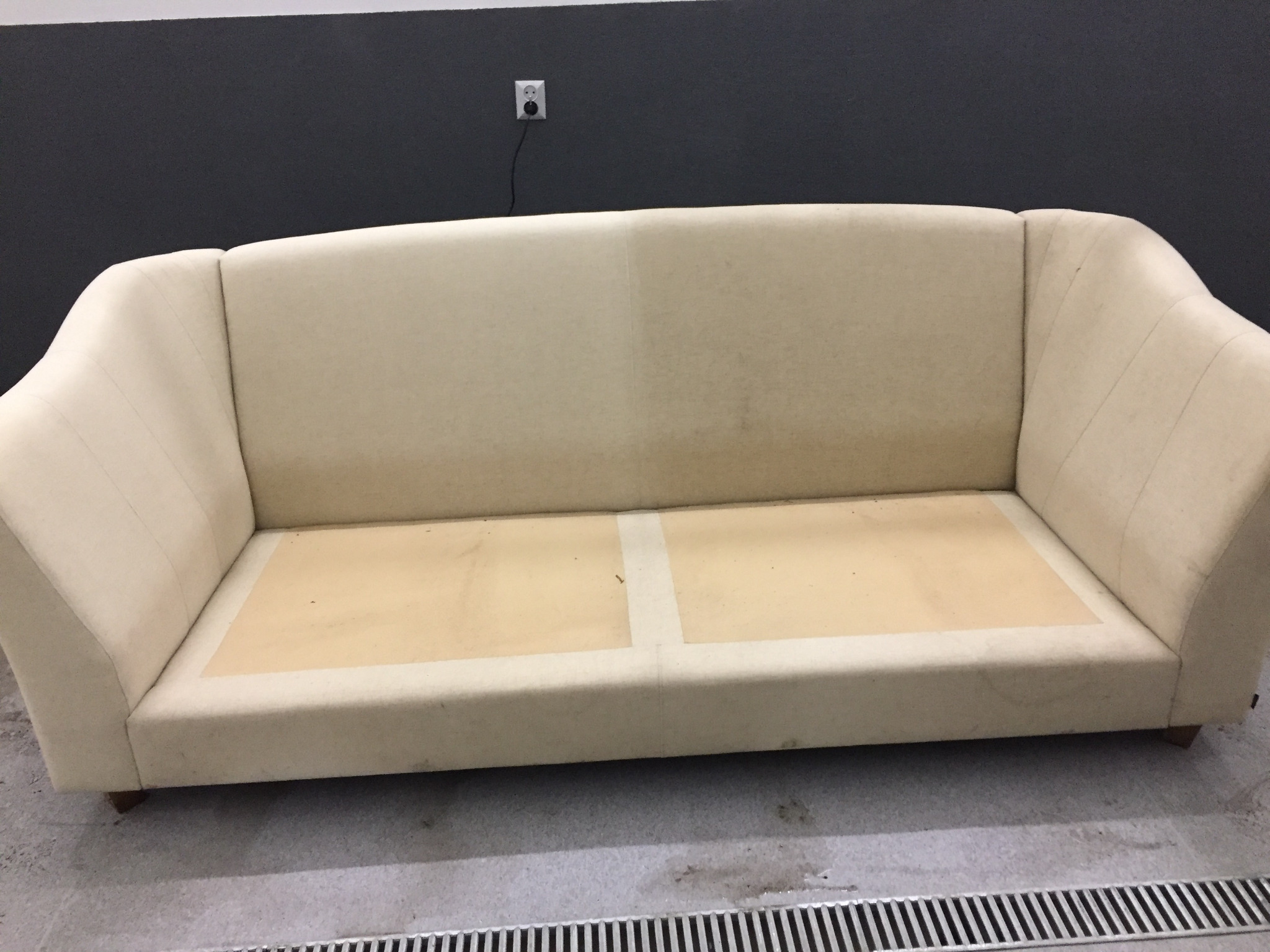 Beżowa kanapa z widocznymi plamami przed czyszczeniem, na szarym tle ściany i kratki ściekowej.