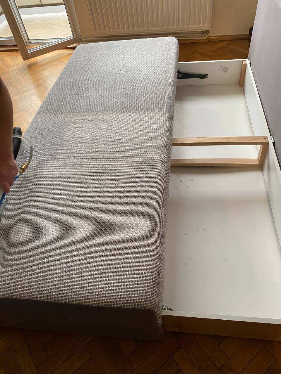 Czyszczenie parowe szarej, rozkładanej sofy z otwartą skrzynią na pościel, widoczna dysza parowa i drewniana podłoga.