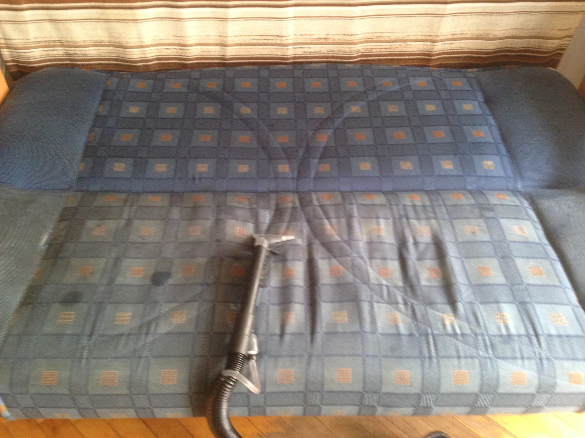 Dysza odkurzacza piorącego na częściowo wyczyszczonej, wzorzystej kanapie w odcieniach niebieskiego i brązu. Górna część kanapy jest jeszcze brudna, a dolna po czyszczeniu jest wyraźnie jaśniejsza.