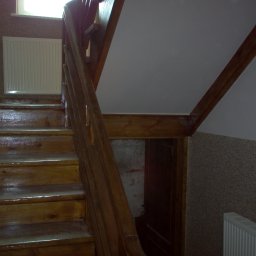 Keu - Drewniane schody z poręczą, z widocznym grzejnikiem panelowym na ścianie i wejściem pod schodami