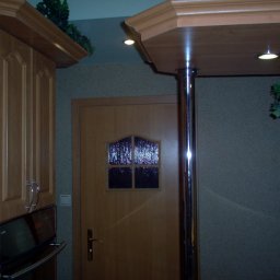 Keu - Fragment kuchni w Toruniu z drewnianymi szafkami, piekarnikiem Amica, drzwiami z szybką i metalową podporą blatu z oświetleniem punktowym.