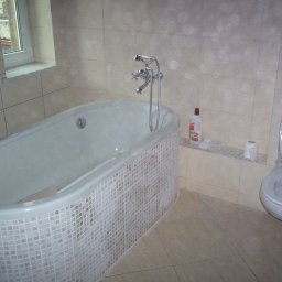 Keu - Ukończona łazienka z owalną wanną obłożoną mozaiką, toaletą zabezpieczoną folią ochronną i płytkami w odcieniach beżu na ścianach i podłodze.