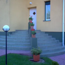 Tarasowe schody wejściowe do domu z ceramiczną okładziną w odcieniach zieleni, ozdobione donicami z czerwonymi kwiatami, oświetlone kulistą lampą.