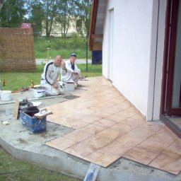 Keu - Dwaj mężczyźni w roboczych ubraniach układają brązowe płytki na betonowym tarasie przed wejściem do budynku, widoczne narzędzia i materiały budowlane.