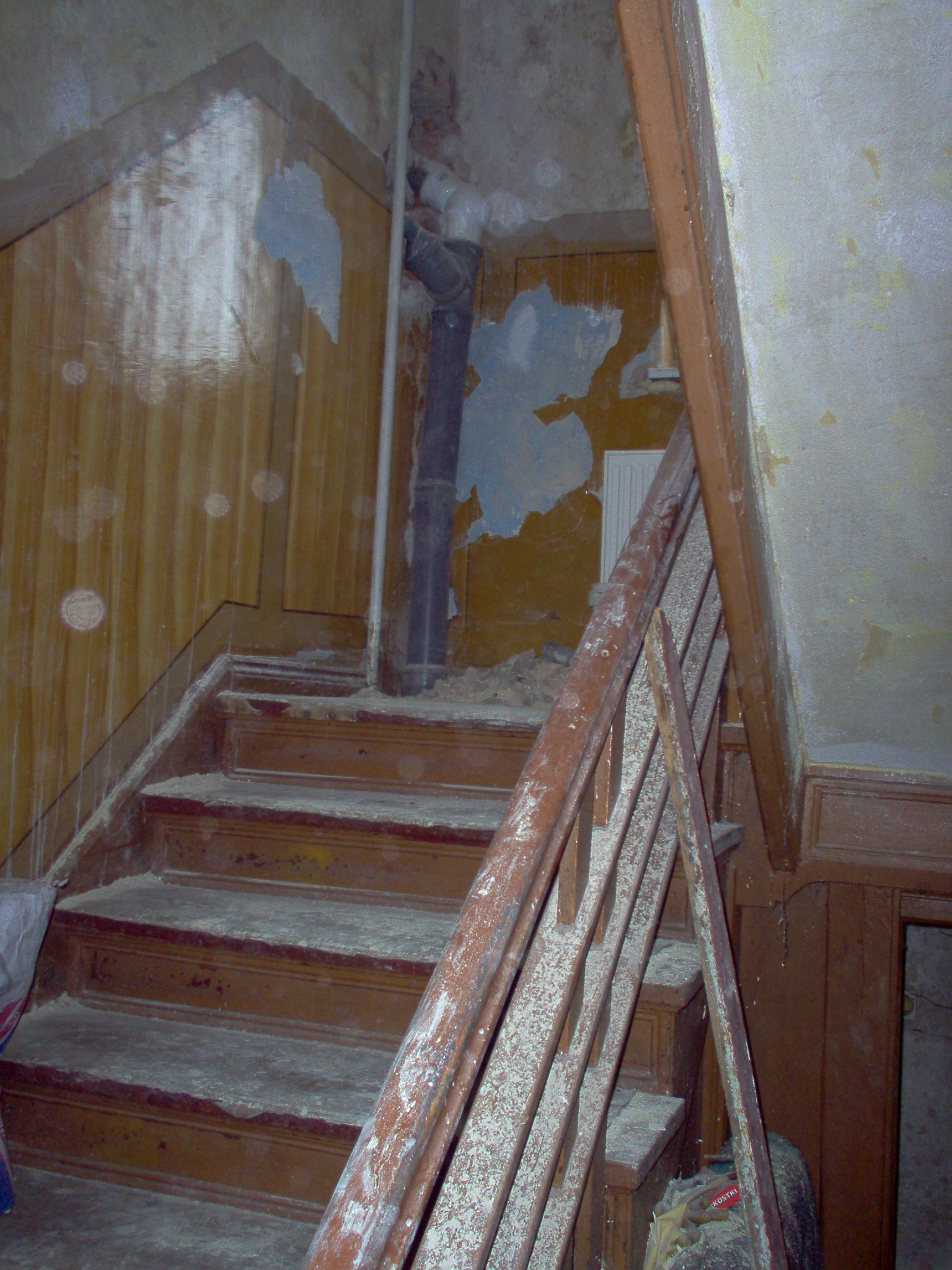 Schody drewniane w trakcie remontu, pokryte grubą warstwą pyłu, z widocznymi fragmentami odchodzącej farby i uszkodzeniami na ścianach.