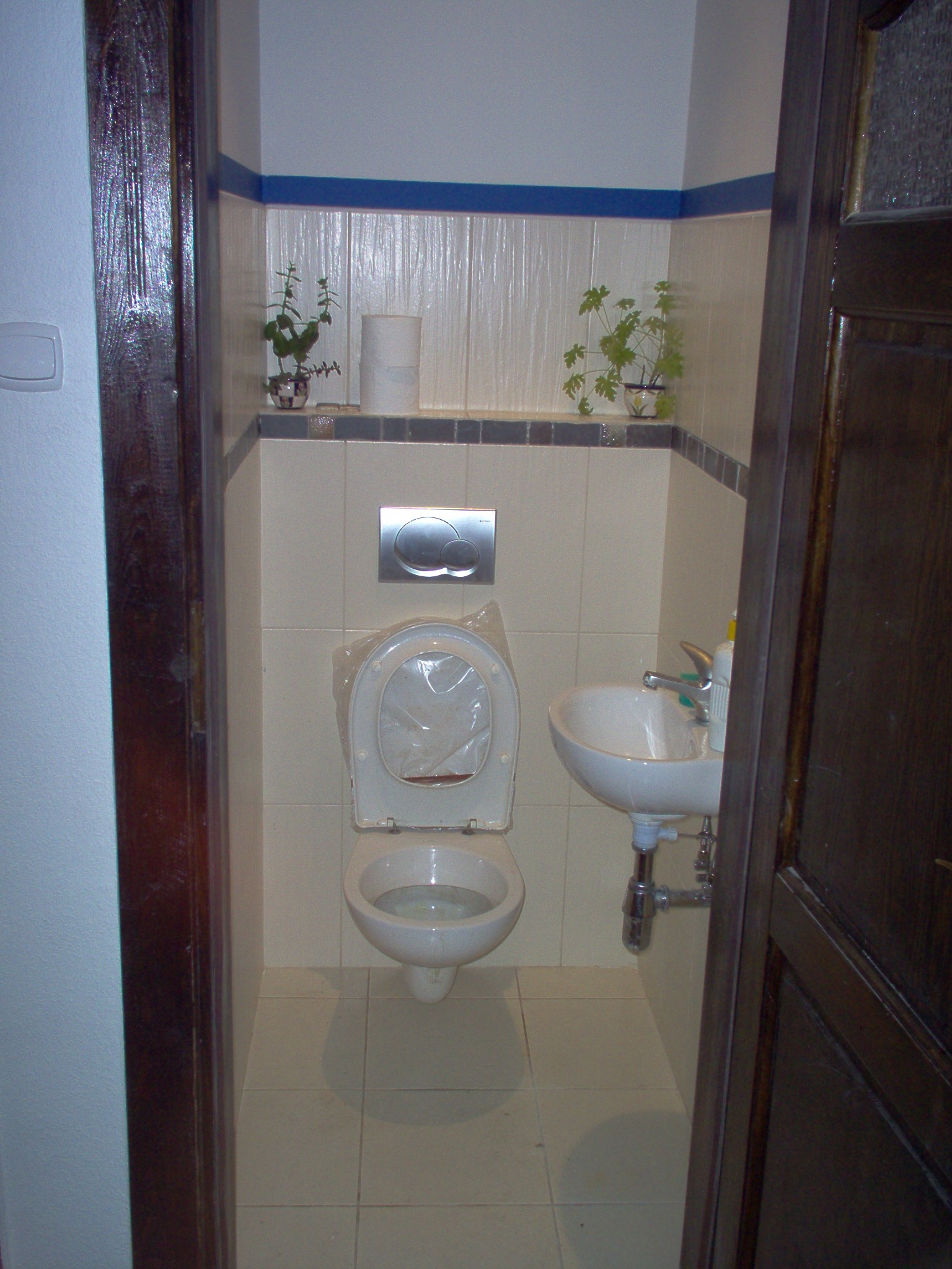 Wnętrze łazienki z toaletą z założoną folią ochronną na desce, umywalką i płytkami w odcieniach beżu, widok zza ciemnych drewnianych drzwi.