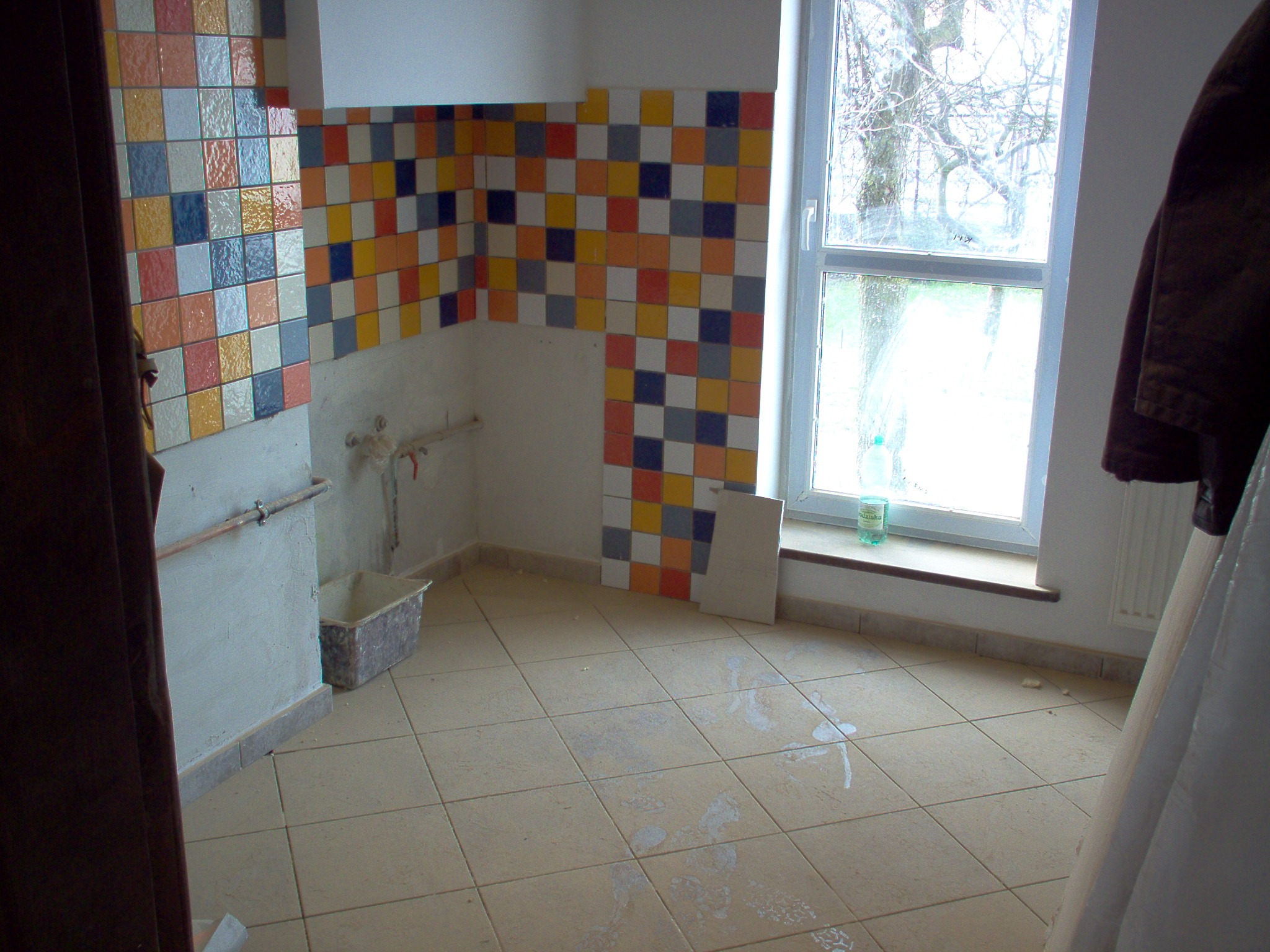 Wnętrze łazienki w trakcie remontu: częściowo ułożone kolorowe płytki na ścianie, podłoga w trakcie kładzenia, widoczne rury i okno z widokiem na drzewo.