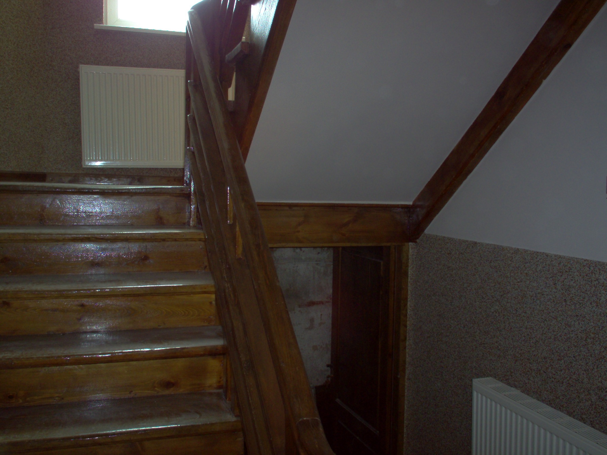 Drewniane schody z poręczą, z widocznym grzejnikiem panelowym na ścianie i wejściem pod schodami