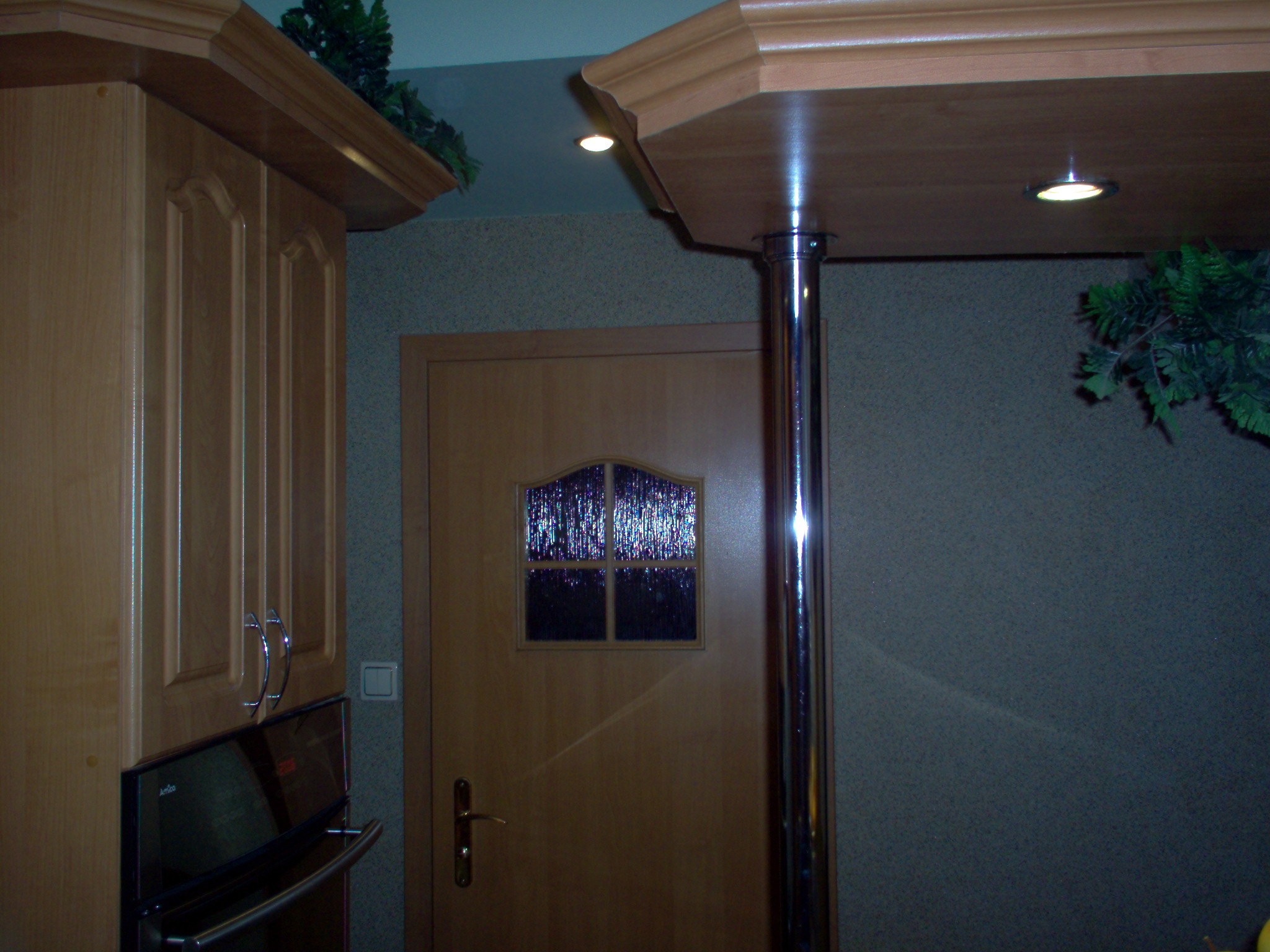 Fragment kuchni w Toruniu z drewnianymi szafkami, piekarnikiem Amica, drzwiami z szybką i metalową podporą blatu z oświetleniem punktowym.