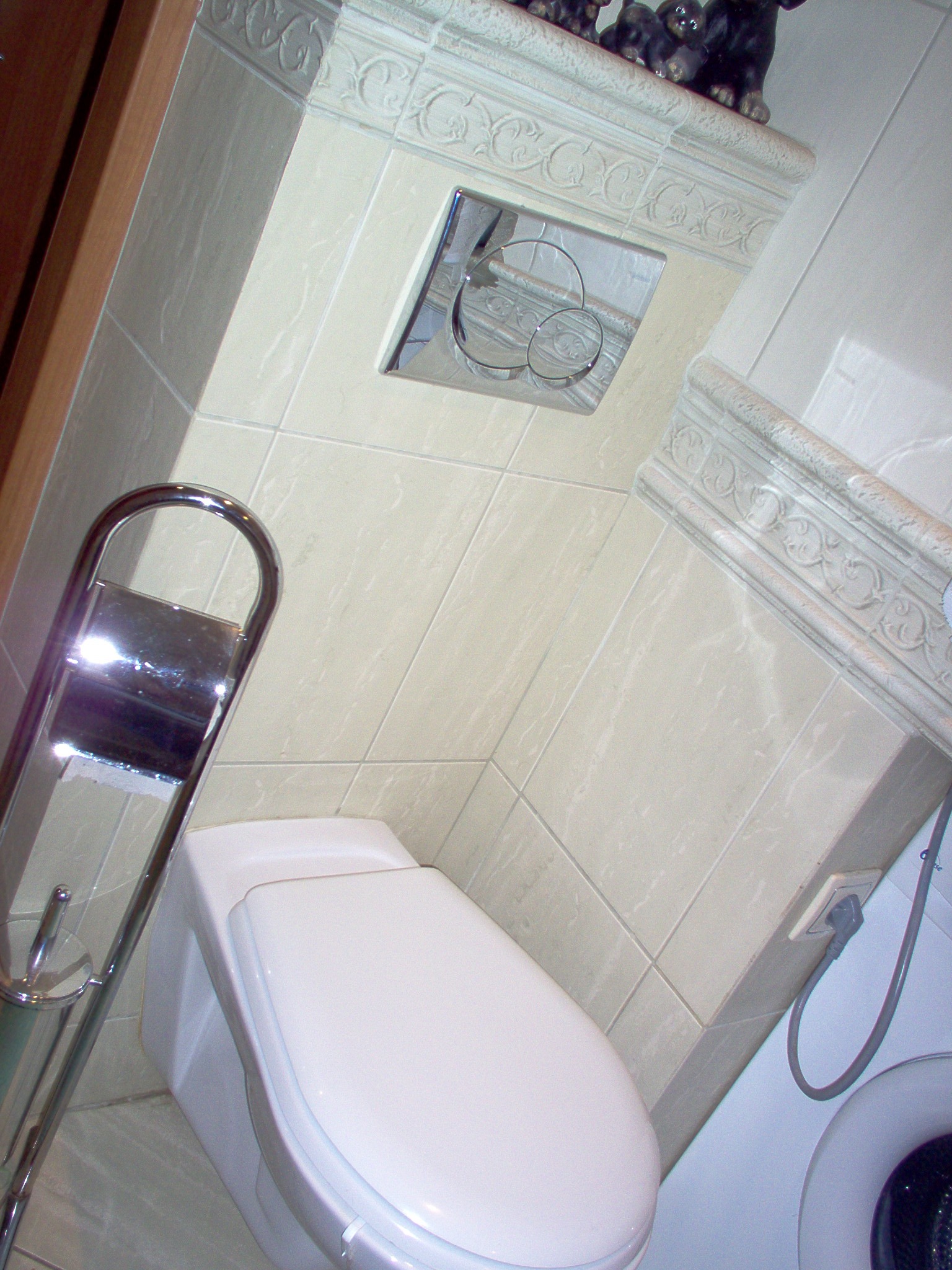 Biała toaleta podwieszana w łazience z beżowymi płytkami, chromowanym przyciskiem spłukiwania i uchwytem na papier toaletowy, widoczny fragment pralki i gniazdko elektryczne.