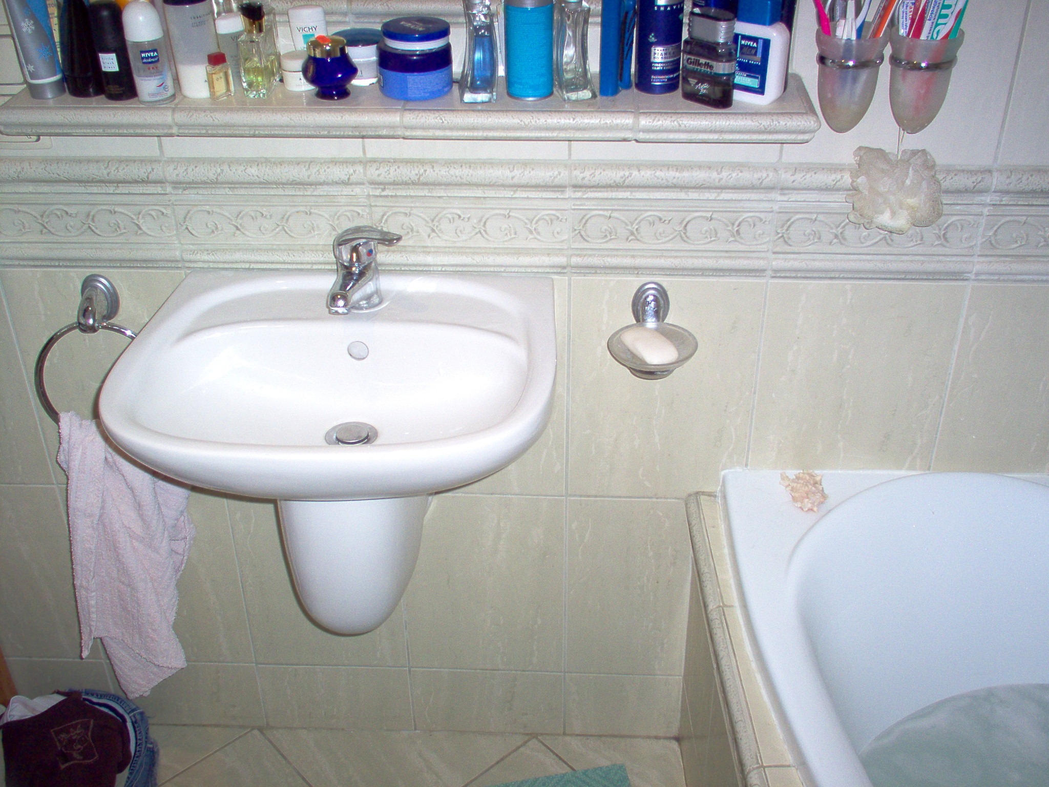 Umywalka ceramiczna ze srebrną baterią w łazience z beżowymi płytkami, mydło w mydelniczce, ręcznik na wieszaku, półka z kosmetykami nad umywalką, fragment wanny w prawym dolnym rogu.