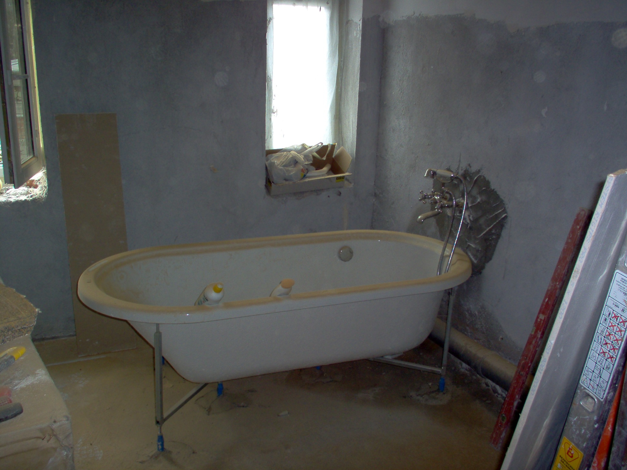 Wolnostojąca wanna na metalowych nogach z regulacją wysokości, ustawiona w surowym pomieszczeniu podczas remontu łazienki, widoczne narzędzia i materiały budowlane.