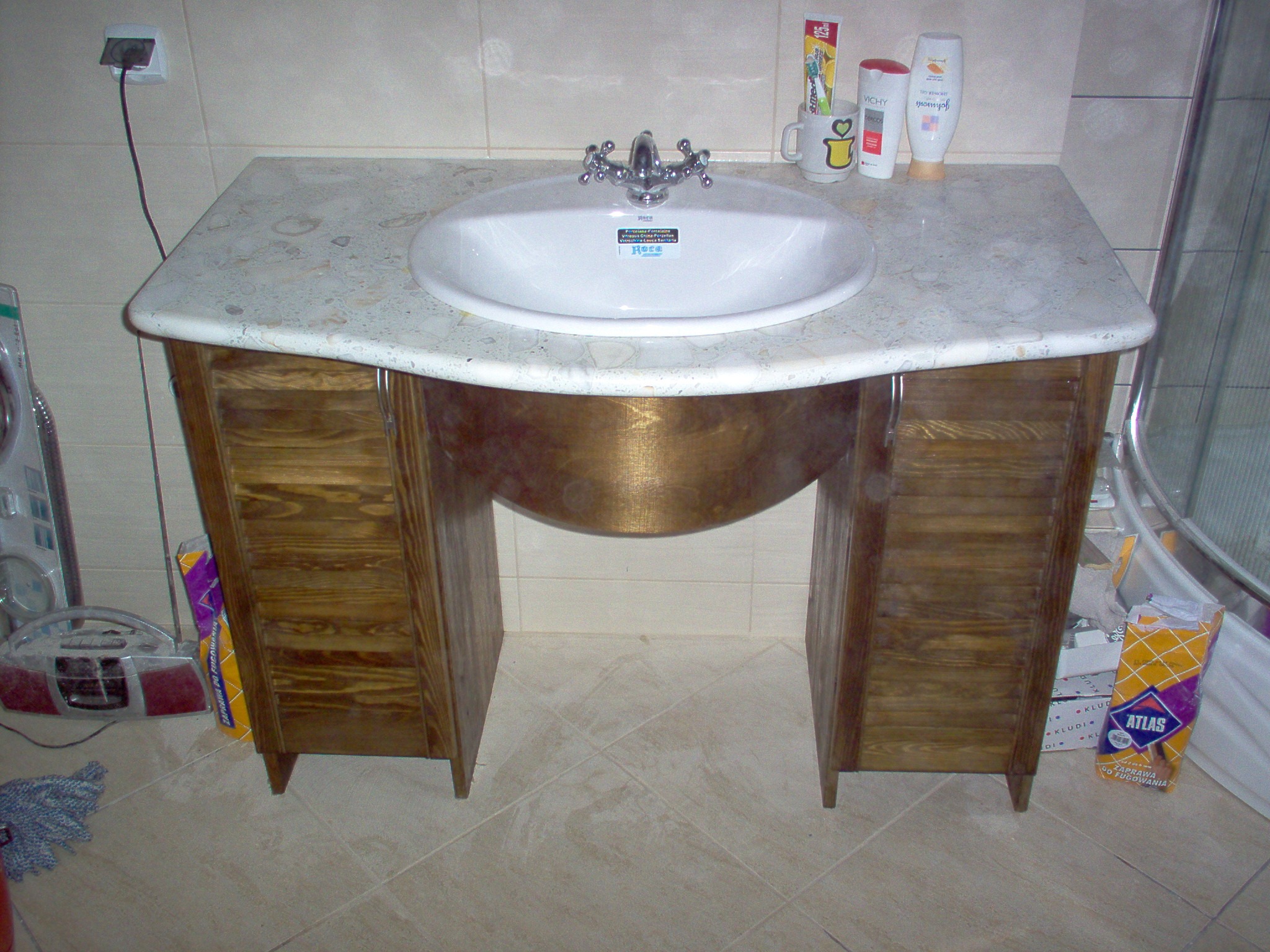 Umywalka nablatowa Roca z chromowaną armaturą na drewnianej szafce z marmurowym blatem, w tle płytki ceramiczne i akcesoria łazienkowe, widoczna zaprawa do fugowania Atlas.