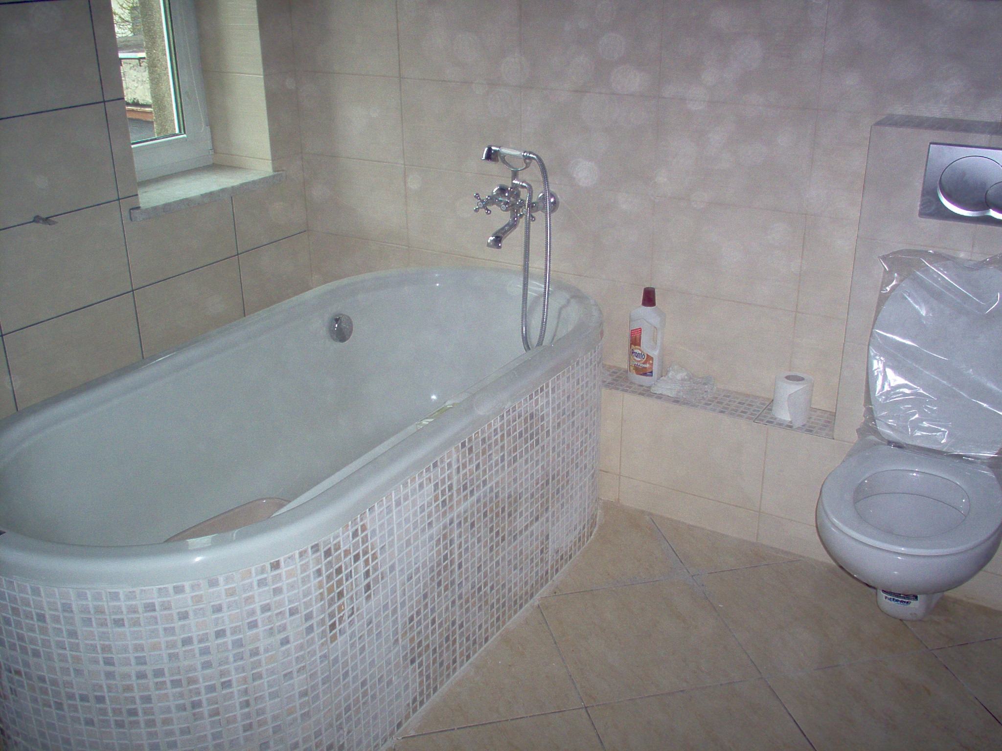 Ukończona łazienka z owalną wanną obłożoną mozaiką, toaletą zabezpieczoną folią ochronną i płytkami w odcieniach beżu na ścianach i podłodze.