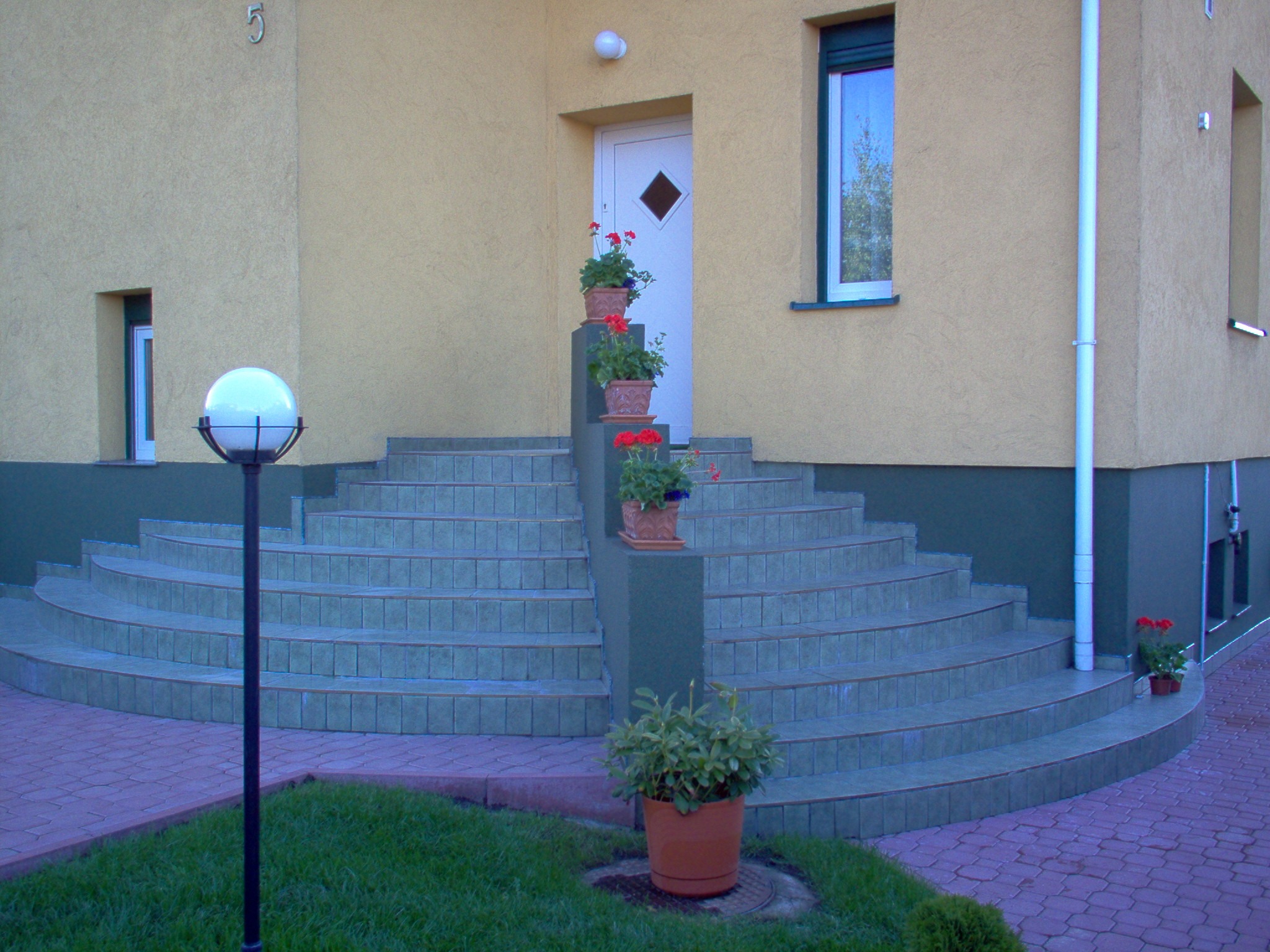 Tarasowe schody wejściowe do domu z ceramiczną okładziną w odcieniach zieleni, ozdobione donicami z czerwonymi kwiatami, oświetlone kulistą lampą.