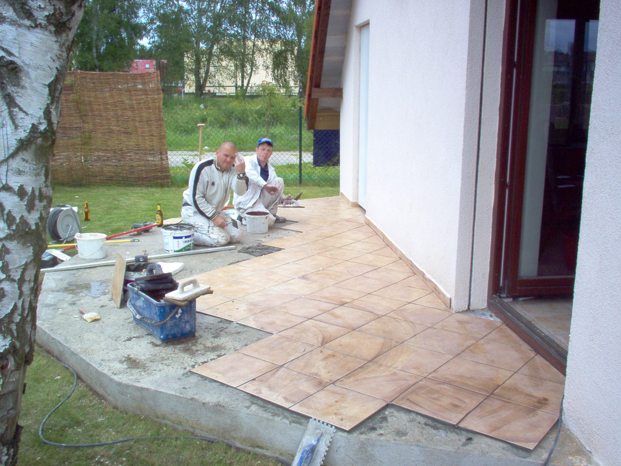 Dwaj mężczyźni w roboczych ubraniach układają brązowe płytki na betonowym tarasie przed wejściem do budynku, widoczne narzędzia i materiały budowlane.