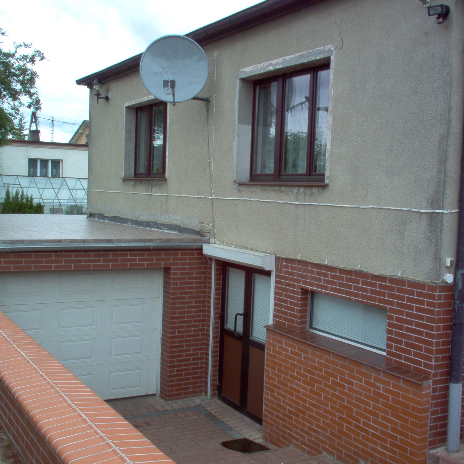 Dwupiętrowy budynek z szarego tynku i ceglaną dobudówką, widoczna satelita na ścianie, biała brama garażowa i brązowe okna, brak estetycznego wykończenia.
