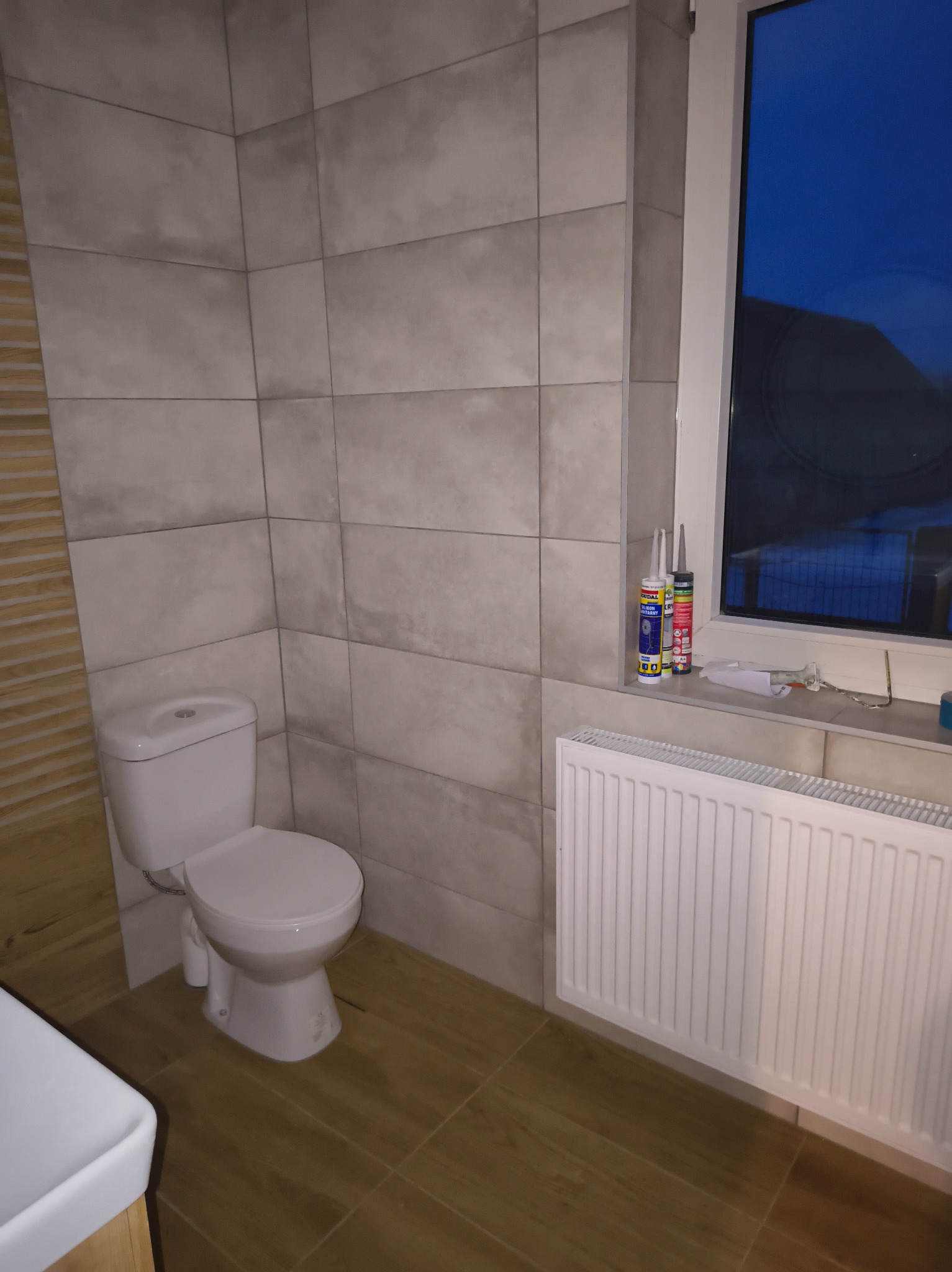 Wykończona łazienka w Wilczogórze: beżowe płytki, biała ceramika i grzejnik. Widoczne tuby silikonu na parapecie. Nowoczesny design.