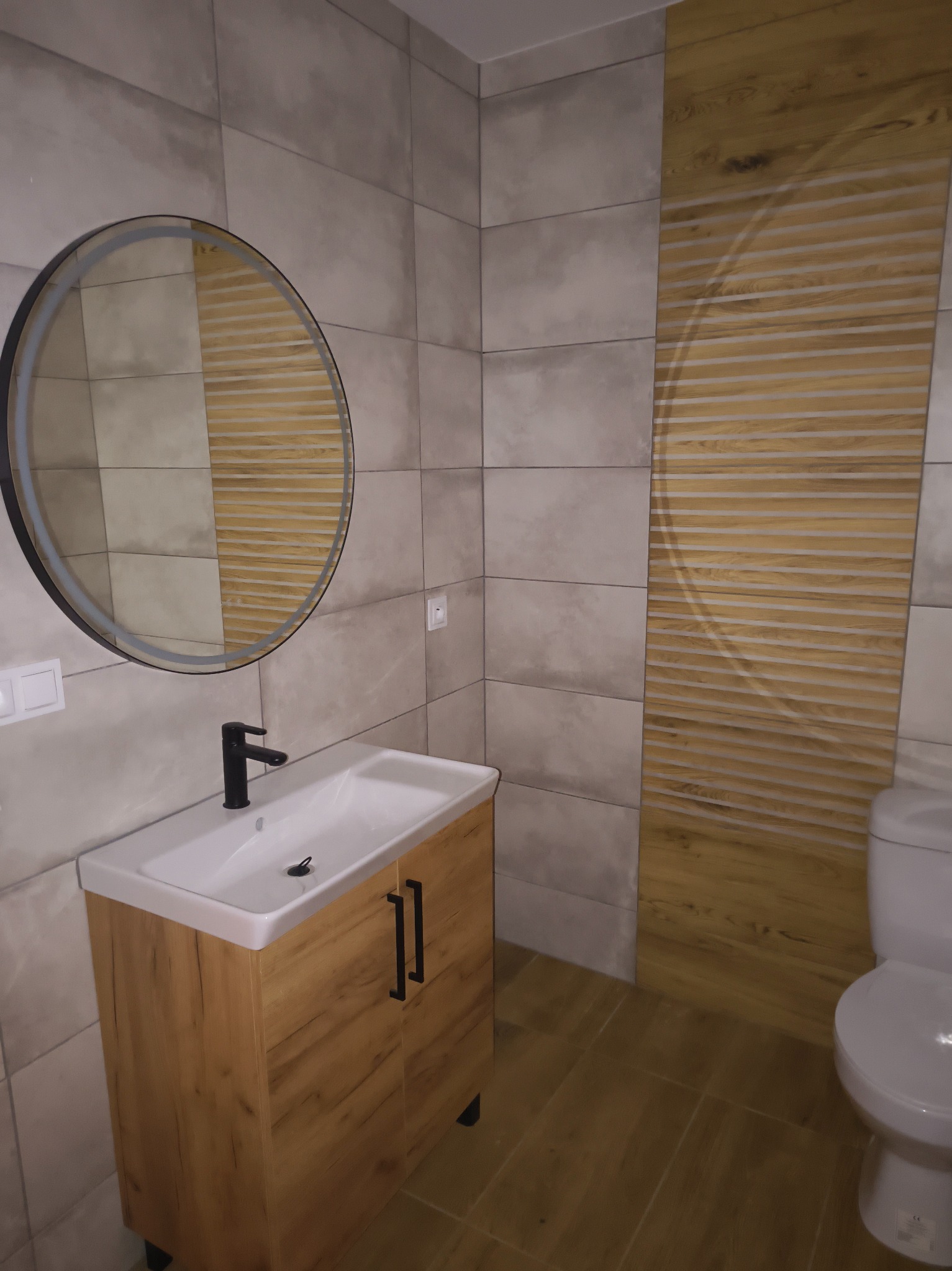 Nowoczesna łazienka z okrągłym lustrem LED nad szafką z umywalką, płytki w odcieniach szarości i drewnopodobne panele na ścianie. Minimalistyczny design.