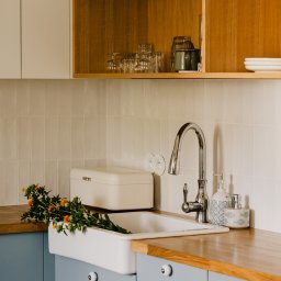 VEGITO ARTUR RUMIŃSKI - Nowoczesna kuchnia z niebieskimi szafkami, drewnianym blatem i białym zlewem farmerskim, ozdobiona bukietem pomarańczowych kwiatów i stylowymi akcesoriami.