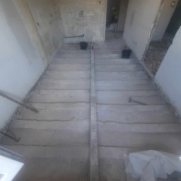 VEGITO ARTUR RUMIŃSKI - Surowa podłoga w trakcie prac przygotowawczych, widoczne ślady po wylewce, narzędzia i wiaderka, wnętrze w stanie deweloperskim.