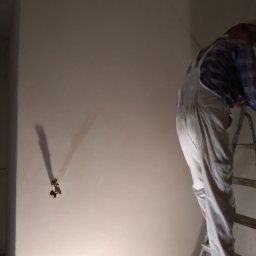 VEGITO ARTUR RUMIŃSKI - Pracownik w kombinezonie na drabinie, wygładza świeżo nałożony tynk na ścianie, widoczne pozostałości po instalacji elektrycznej.