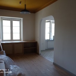 VEGITO ARTUR RUMIŃSKI - Wnętrze po remoncie z drewnianą podłogą, świeżo pomalowanymi ścianami w jasnym odcieniu i żółtym sufitem, częściowo zakryte meble folią ochronną.