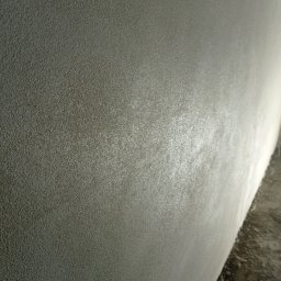 VEGITO ARTUR RUMIŃSKI - Szara ściana z wyraźną, drobną fakturą, sfotografowana pod kątem, z widocznym fragmentem podłogi w dolnym rogu. Na powierzchni ściany widoczne delikatne refleksy światła.