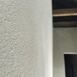 VEGITO ARTUR RUMIŃSKI - Szara ściana z wyraźną, ziarnistą fakturą, widoczna krawędź ściany, fragment sufitu z drewnianymi belkami i zarys otworu drzwiowego w tle