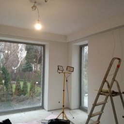VEGITO ARTUR RUMIŃSKI - Pokój w trakcie prac wykończeniowych; widoczne świeżo pomalowane ściany, aluminiowa drabina oparta o ścianę z wiszącymi kablami oraz reflektor na statywie oświetlający wnętrze. Duże okno z widokiem...