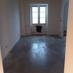 VEGITO ARTUR RUMIŃSKI - Szara, mokra wylewka anhydrytowa w pustym pokoju z oknem. Widoczne wystające kable elektryczne ze ściany. Przy oknie wisi czarny materiał.