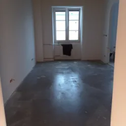 Szara, mokra wylewka anhydrytowa w pustym pokoju z oknem. Widoczne wystające kable elektryczne ze ściany. Przy oknie wisi czarny materiał.