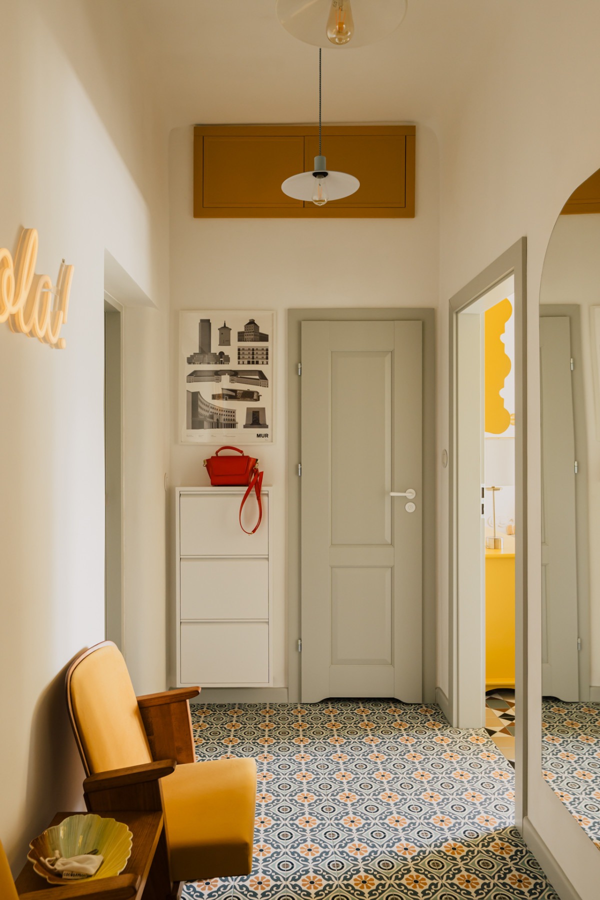 Wnętrze przedpokoju w stylu hiszpańskim z wzorzystą podłogą, żółtym fotelem i neonowym napisem 'hola!' na ścianie, widoczne drzwi i szafka na buty.