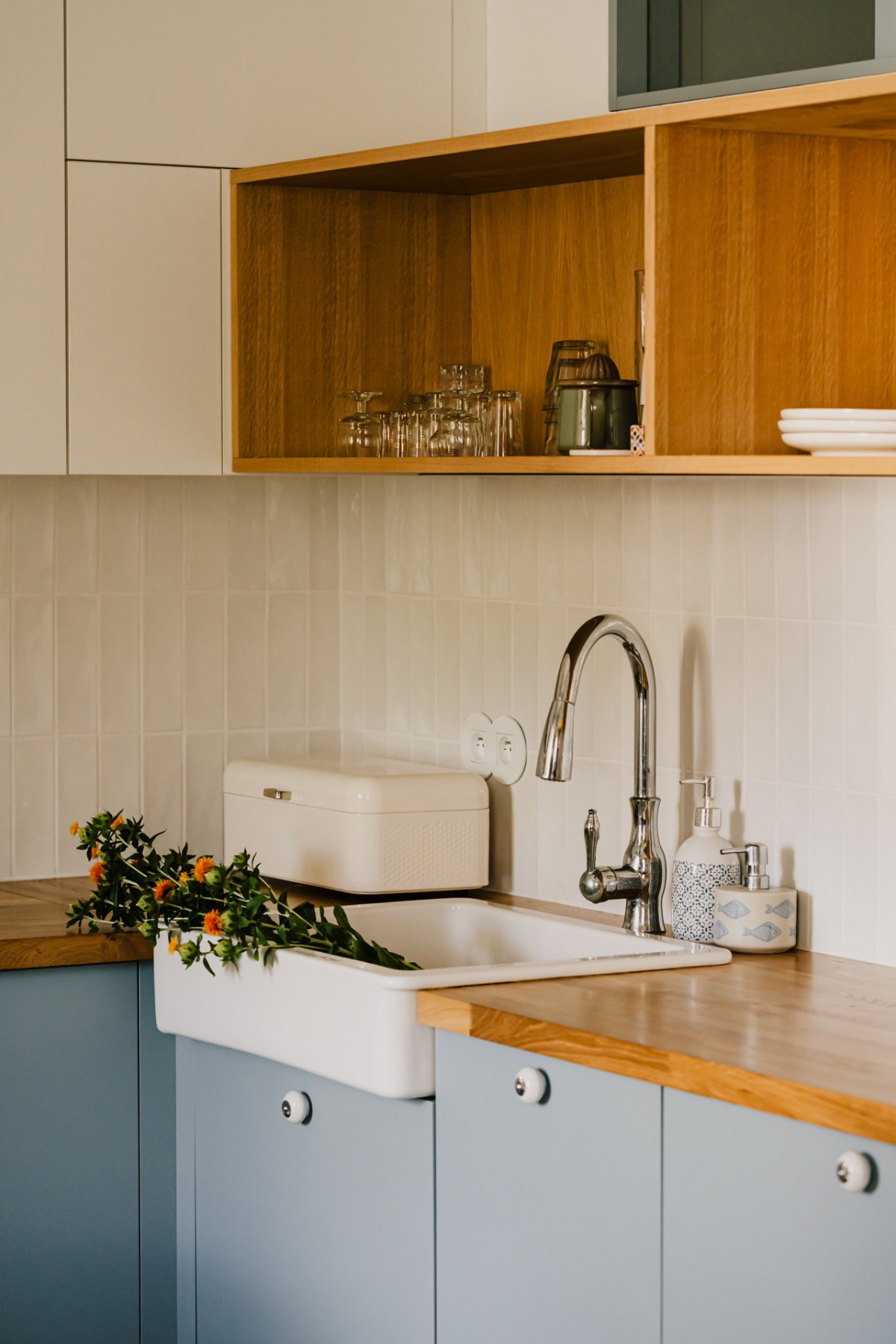 Nowoczesna kuchnia z niebieskimi szafkami, drewnianym blatem i białym zlewem farmerskim, ozdobiona bukietem pomarańczowych kwiatów i stylowymi akcesoriami.