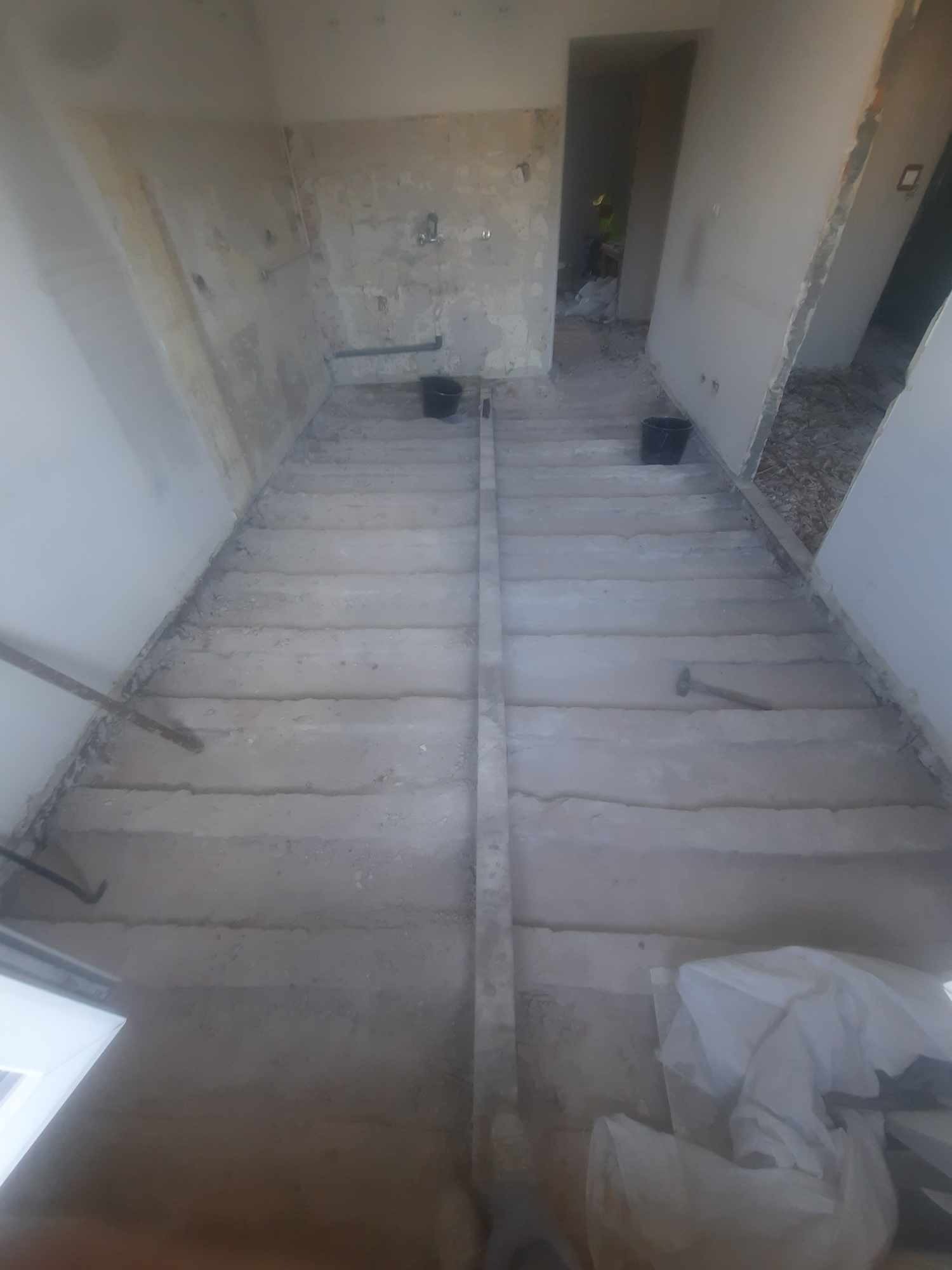 Surowa podłoga w trakcie prac przygotowawczych, widoczne ślady po wylewce, narzędzia i wiaderka, wnętrze w stanie deweloperskim.