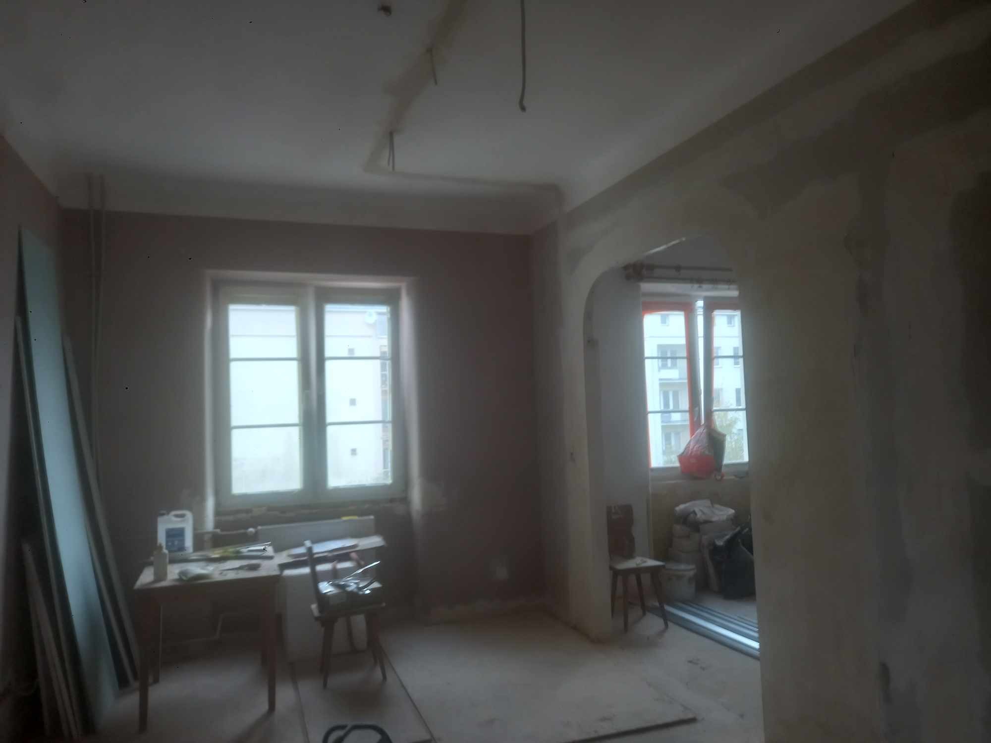 Pokój w trakcie remontu, ściany pokryte świeżym tynkiem, widoczne narzędzia i materiały budowlane na prowizorycznym stole, surowy stan wykończenia, przejście między pokojami w formie łuku.