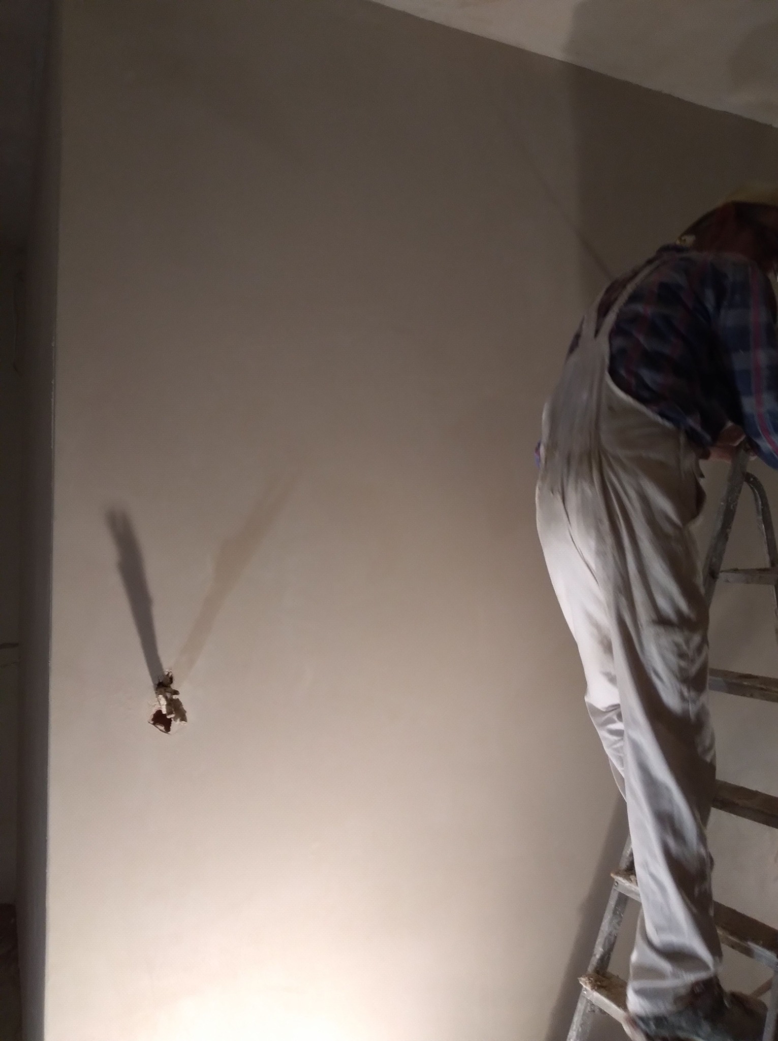 Pracownik w kombinezonie na drabinie, wygładza świeżo nałożony tynk na ścianie, widoczne pozostałości po instalacji elektrycznej.