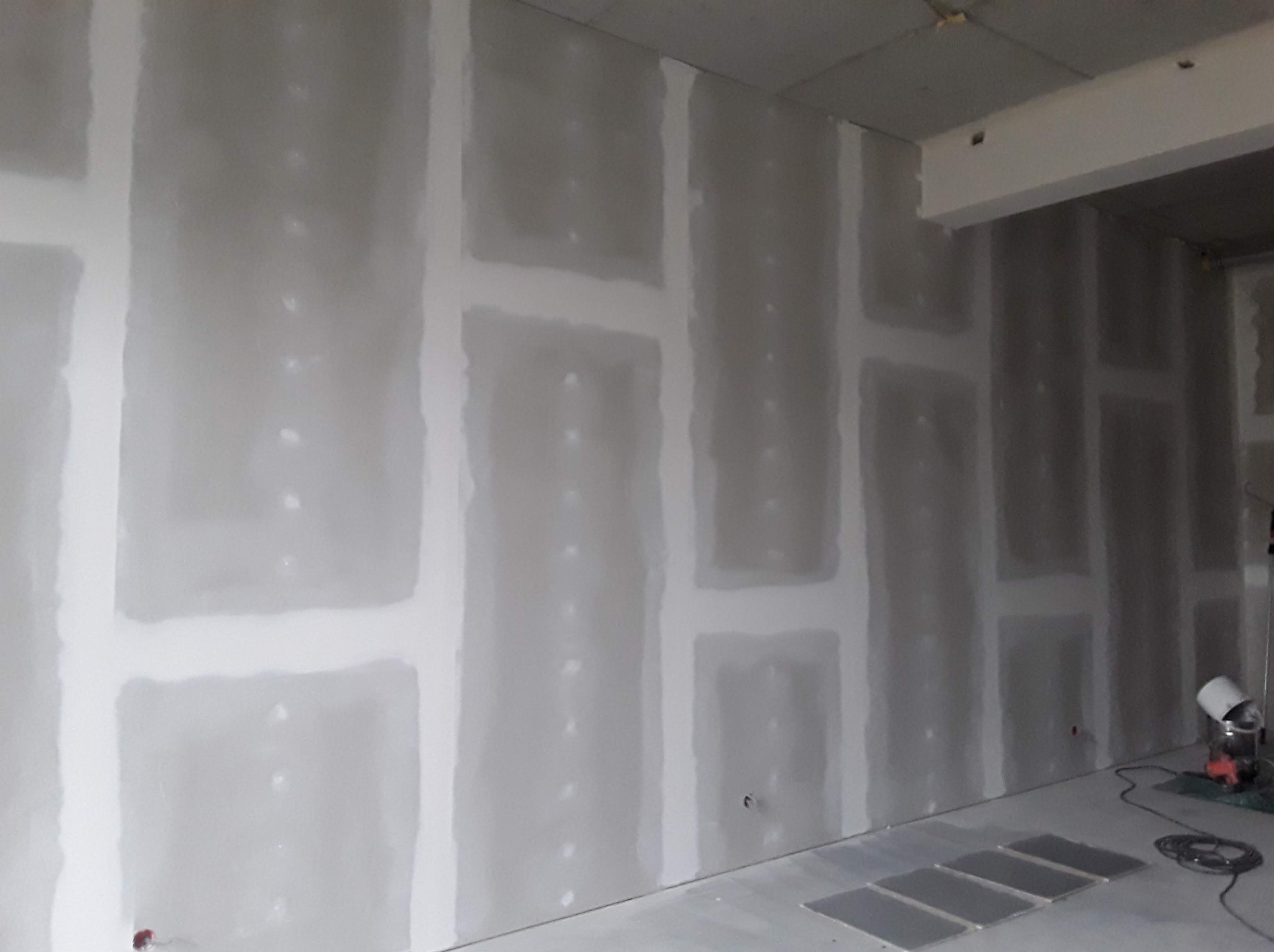 Wnętrze w trakcie remontu: ściany pokryte płytami gipsowo-kartonowymi, widoczne łączenia szpachlowane, fragment sufitu i podłogi, narzędzie do szpachlowania przy ścianie.