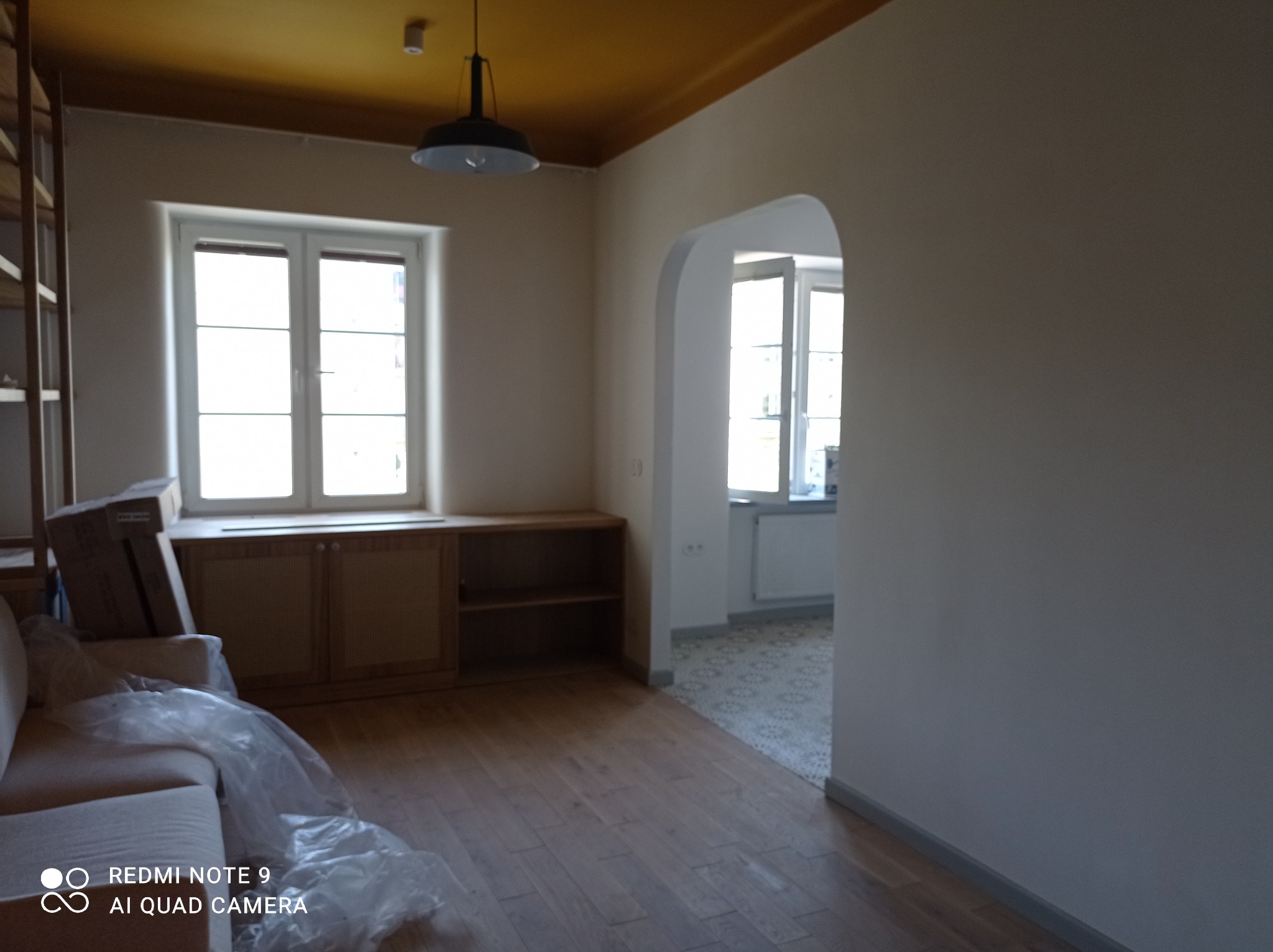 Wnętrze po remoncie z drewnianą podłogą, świeżo pomalowanymi ścianami w jasnym odcieniu i żółtym sufitem, częściowo zakryte meble folią ochronną.