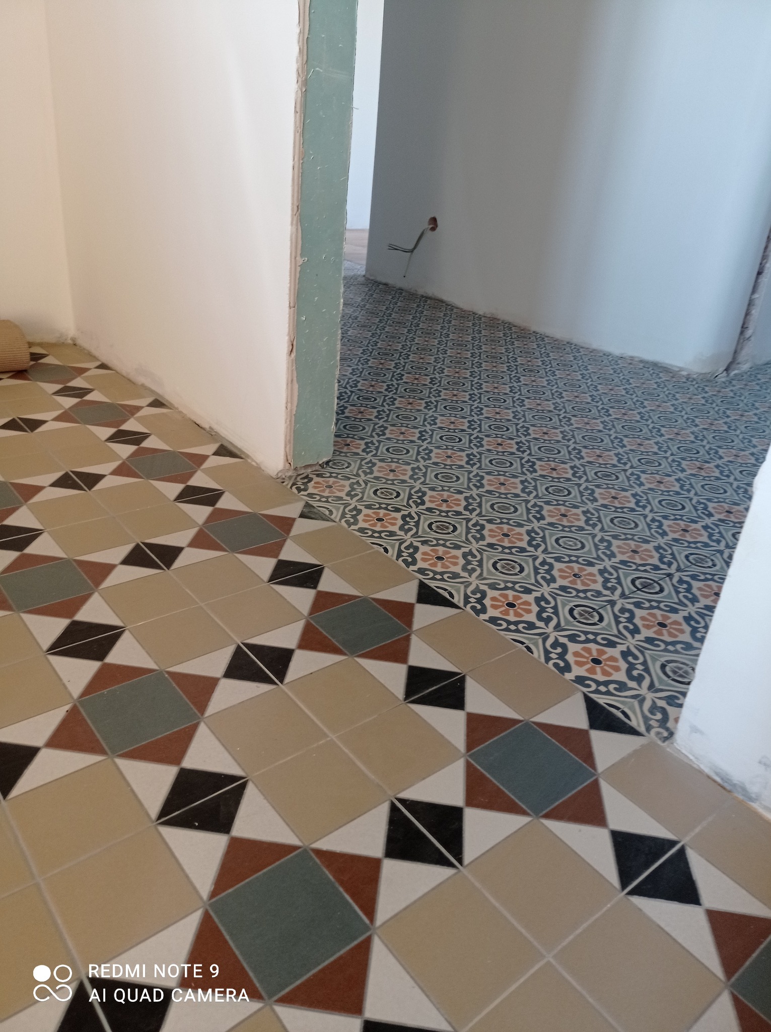 Dwie różne wzorzyste podłogi z płytek ceramicznych w przejściu między pokojami, widoczne niedoskonałości przy łączeniu z ścianą.