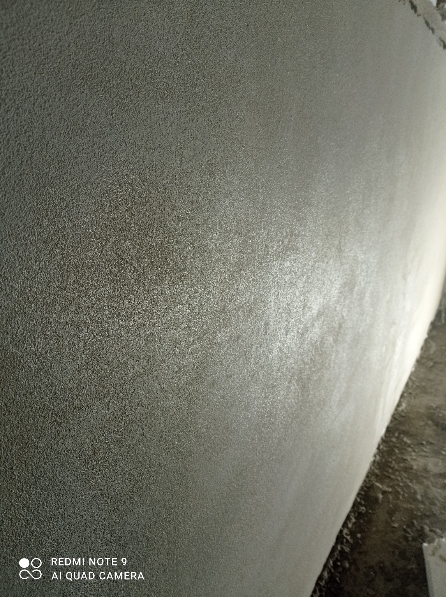 Szara ściana z wyraźną, drobną fakturą, sfotografowana pod kątem, z widocznym fragmentem podłogi w dolnym rogu. Na powierzchni ściany widoczne delikatne refleksy światła.