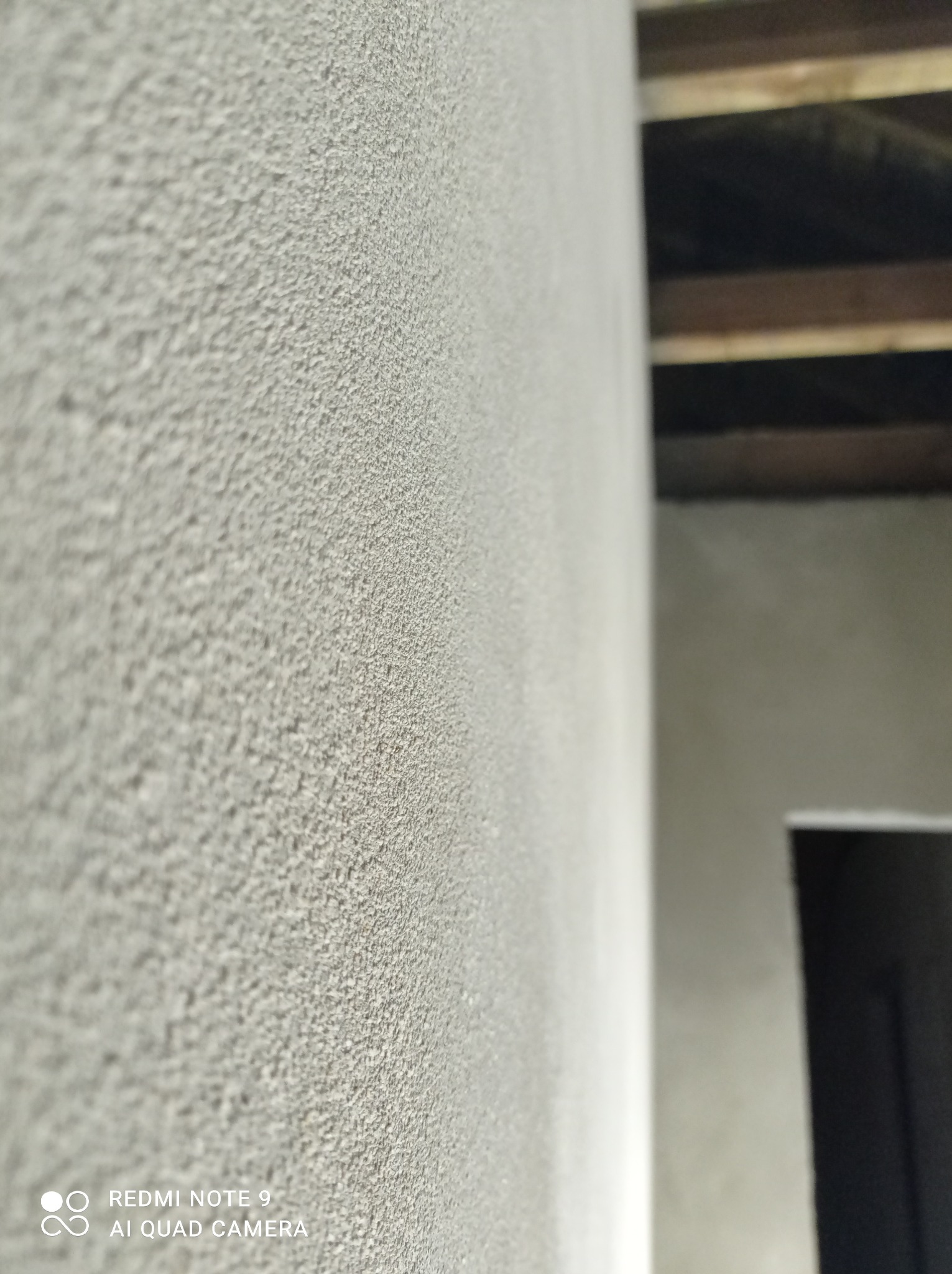 Szara ściana z wyraźną, ziarnistą fakturą, widoczna krawędź ściany, fragment sufitu z drewnianymi belkami i zarys otworu drzwiowego w tle