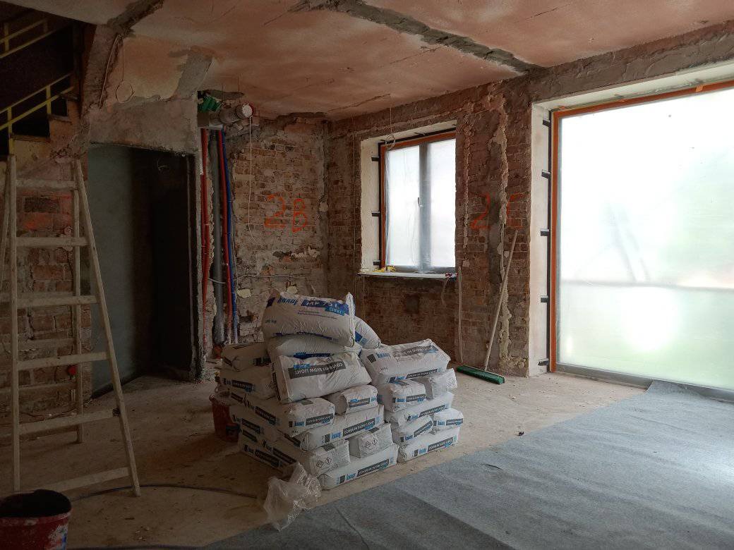 Wnętrze w trakcie remontu: surowe ściany z cegły, zakryte okno, przygotowane worki z zaprawą i częściowo wykonana wylewka na podłodze.