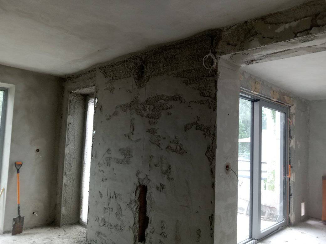 Surowe wnętrze w trakcie prac wykończeniowych, ściany pokryte wstępną warstwą zaprawy, widoczne przewody elektryczne, szpachelka oparta o ścianę i przesuwne drzwi balkonowe.