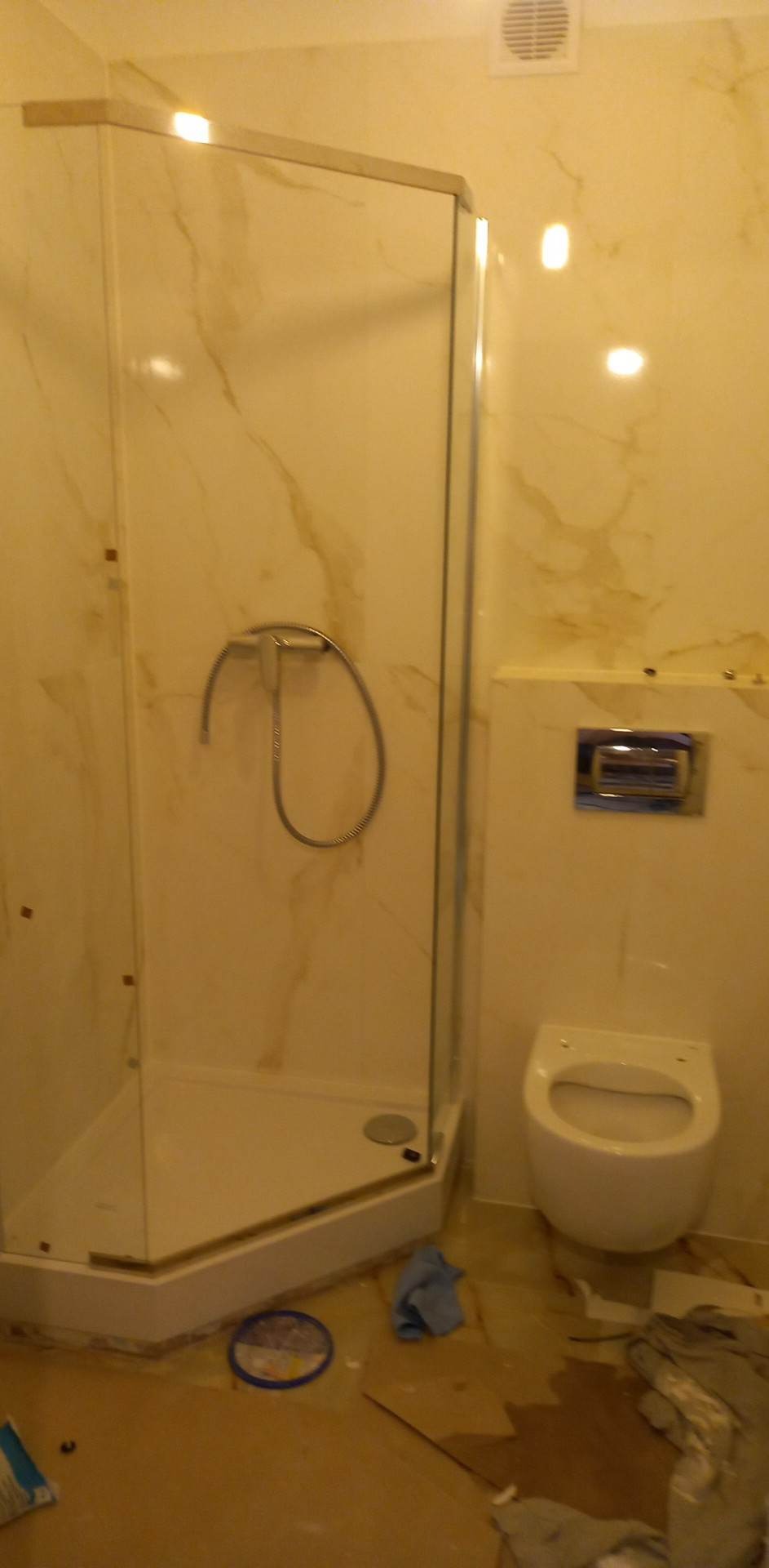 Narożna kabina prysznicowa z zamontowaną słuchawką, wisząca toaleta i marmurowe płytki ścienne w trakcie remontu łazienki, widoczne zabrudzenia na podłodze.