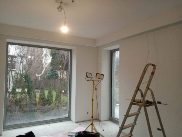 Pokój w trakcie prac wykończeniowych; widoczne świeżo pomalowane ściany, aluminiowa drabina oparta o ścianę z wiszącymi kablami oraz reflektor na statywie oświetlający wnętrze. Duże okno z widokiem...