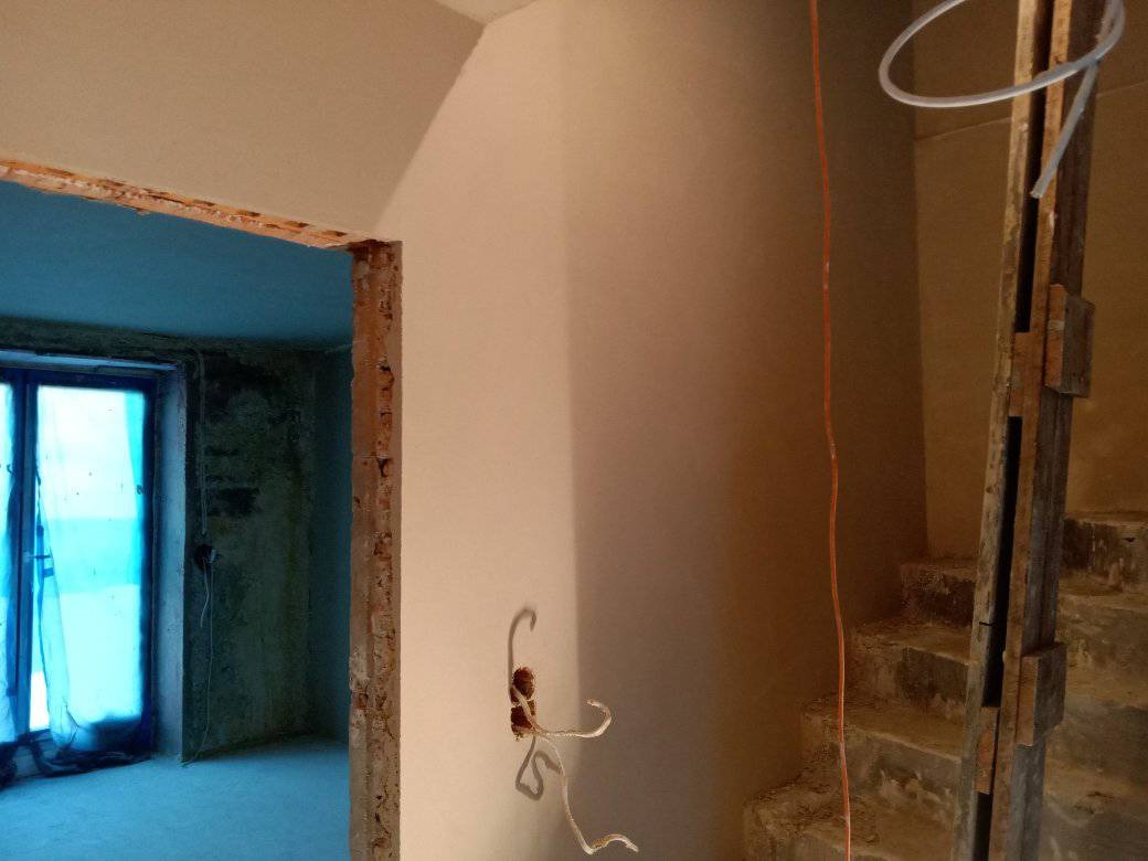 Wnętrze w trakcie prac wykończeniowych: gładkie ściany po tynkowaniu, widoczne przewody elektryczne wystające ze ściany, surowe obramowanie drzwi i fragment schodów w tle.