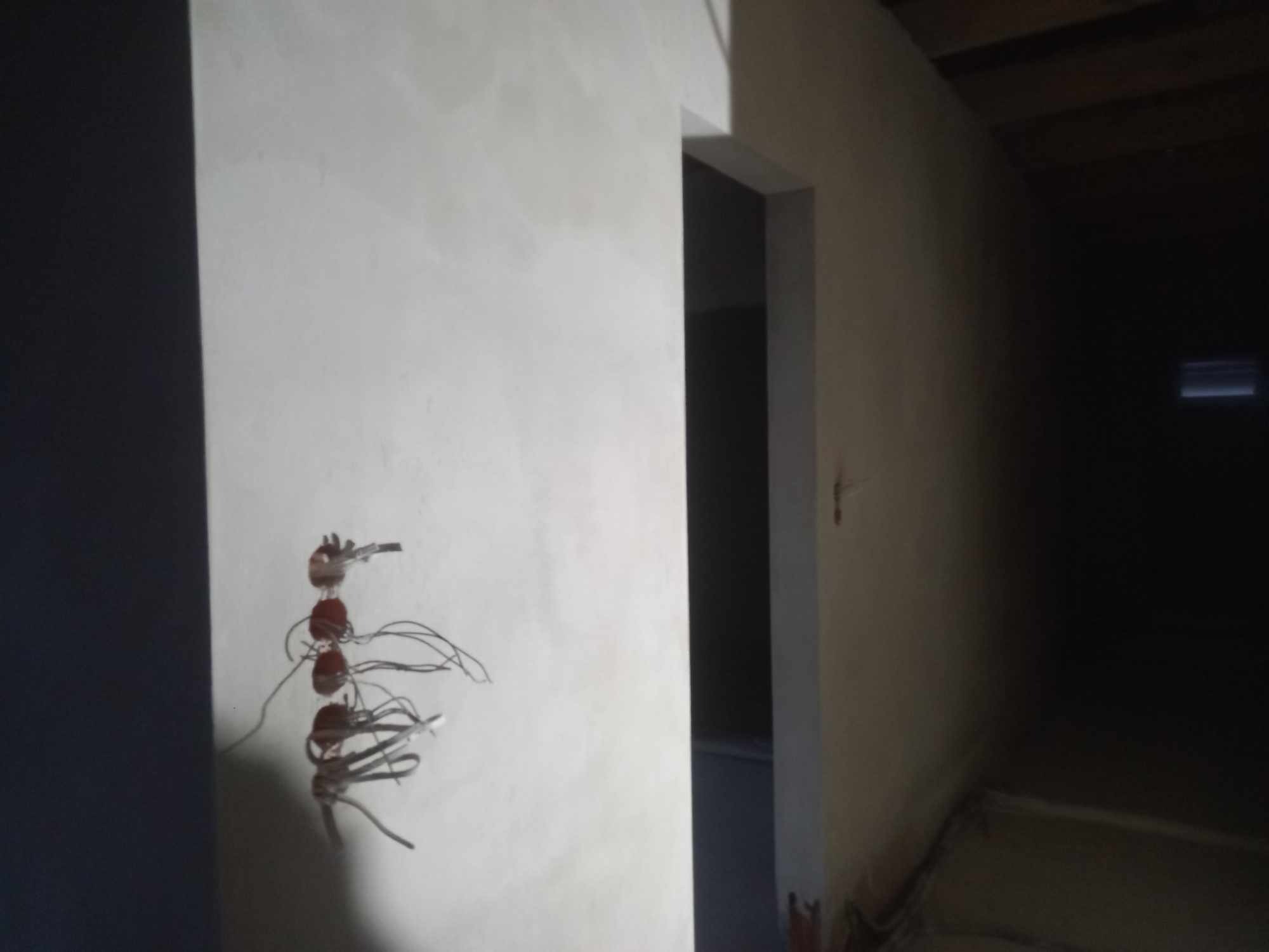 Świeżo otynkowana ściana z wyprowadzonymi kablami elektrycznymi, widok na ciemne pomieszczenie przez otwór drzwiowy.