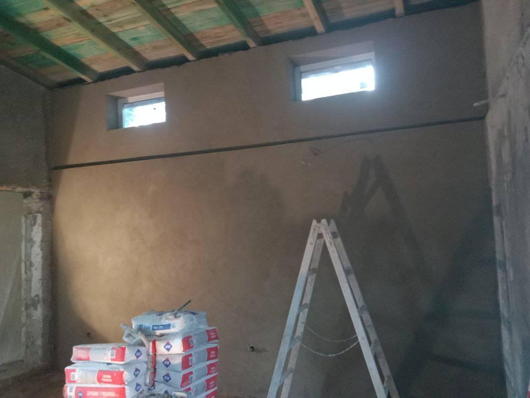 Świeżo nałożony tynk na ścianie wewnętrznej, widoczne dwa małe okna pod sufitem, drabina oparta o ścianę i stos worków z zaprawą tynkarską na podłodze.