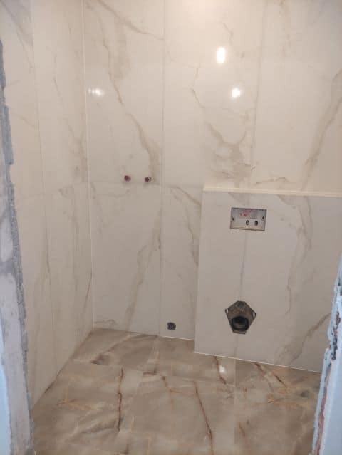 Łazienka w trakcie wykańczania: ściany i podłoga wyłożone płytkami imitującymi marmur, widoczny stelaż podtynkowy WC z otworem na przyłącze, fragment surowej ściany z lewej strony.